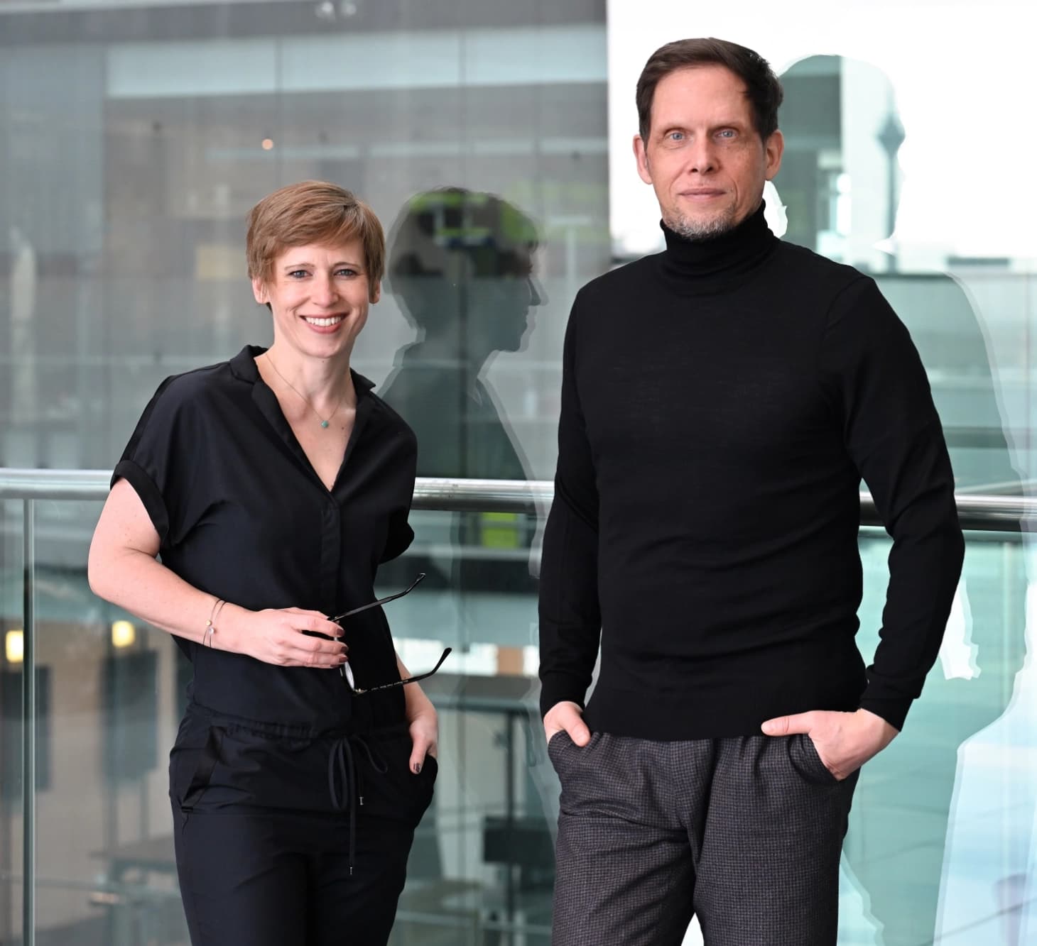 Swantje und Peter stehen vor einer Glasscheibe und schauen in die Kamera